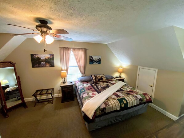 Quarto aconchegante com cama, um ventilador de teto, janela com cortinas, mesa de cabeceira com abajur e uma porta.