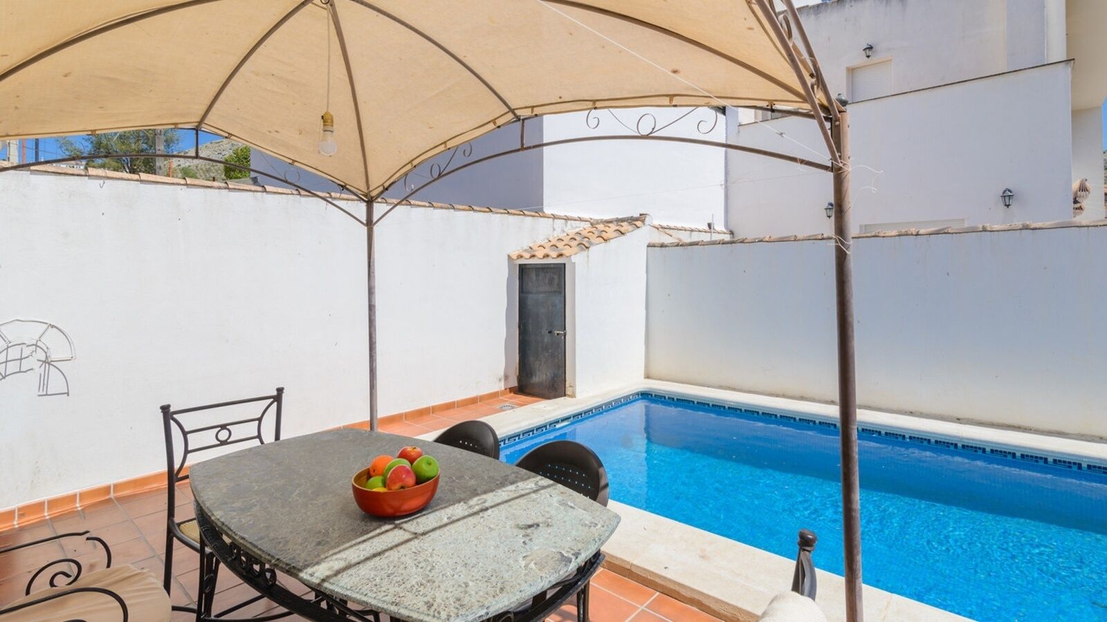 A poolside area with a table and chairs, a bowl of fruit, and a covered seating area.