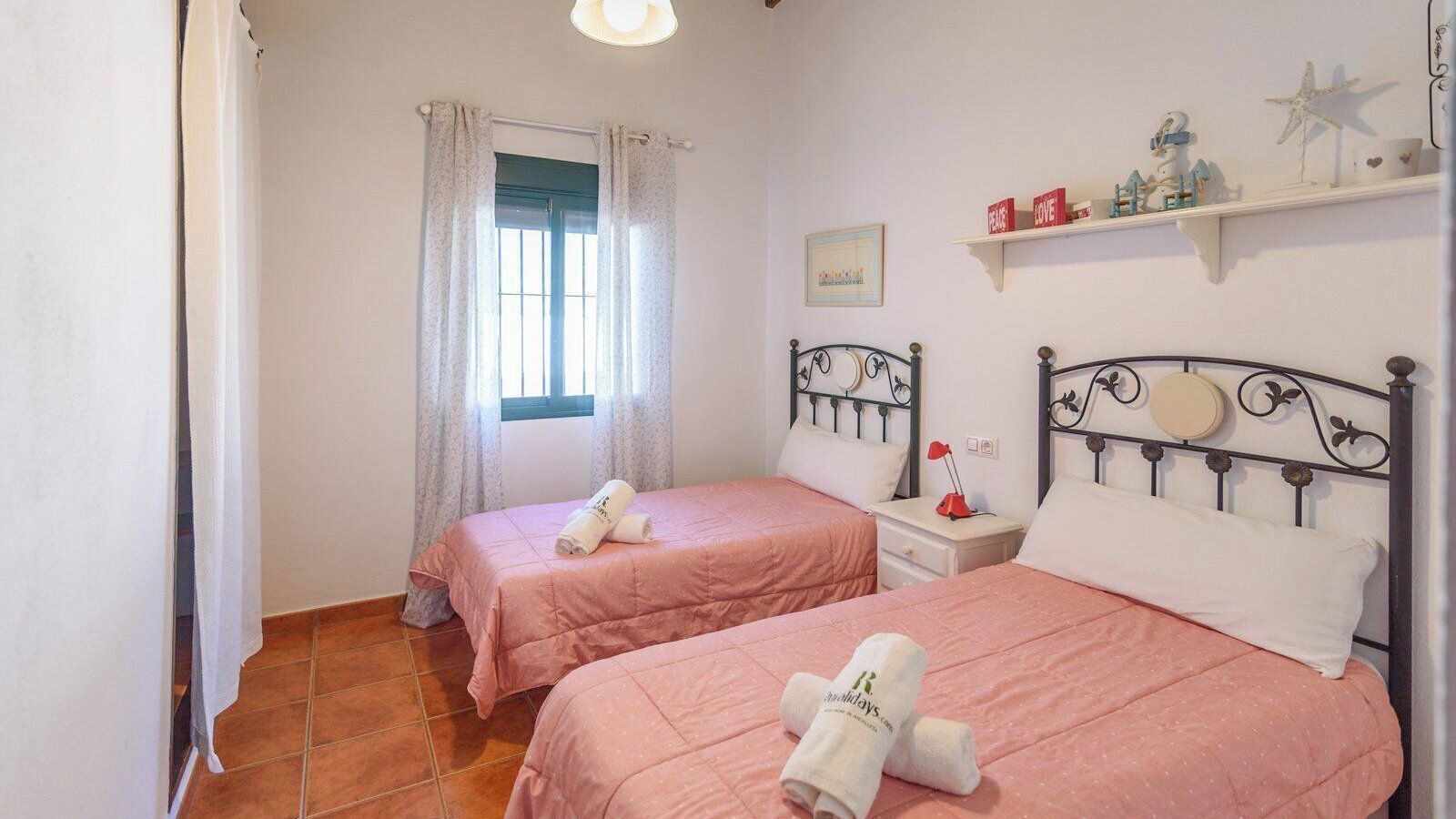 A bedroom with two beds, a window with curtains, a bedside table with a lamp, and a shelf with decorative items.