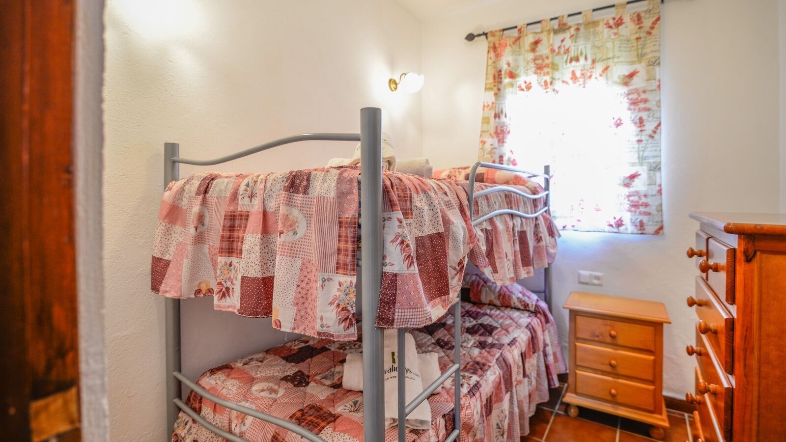Un letto a castello con trapunta floreale, un comò in legno e una finestra con tende a fiori.
