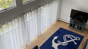 Ein Wohnzimmer mit Sofa, Fernseher, Deckenventilator und großen Fenstern mit transparenten Vorhängen.