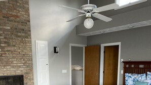 Ein Schlafzimmer mit einem hölzernen Bett, einem Deckenventilator, einem Kamin aus Ziegelsteinen und einem großen Fenster.