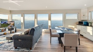 A modern living room with a large sofa, a dining area, and a kitchen. There are large windows offering a view of the sea. - Orca View Suite 10 Mile Point (Victoria)