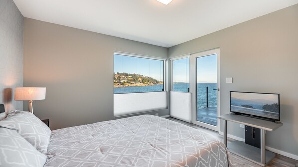 A modern bedroom with a bed, a TV, and a view of a coastal landscape through large windows. - Orca View Suite 10 Mile Point (Victoria)