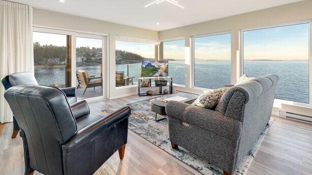 A modern living room with a large window overlooking a body of water, a leather sofa, a black armchair, and a small table with books and decorative items. - Orca View Suite 10 Mile Point (Victoria)