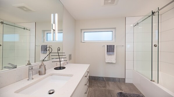 A modern bathroom with a large mirror, dual sinks, a glass shower enclosure, and a small window providing natural light.
