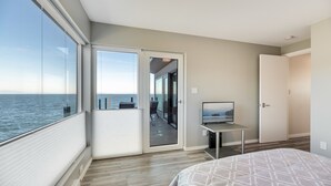 A modern bedroom with a large window, a balcony with a view of the sea, a flat-screen TV, and a bed with a patterned cover. - Orca View Suite 10 Mile Point (Victoria)