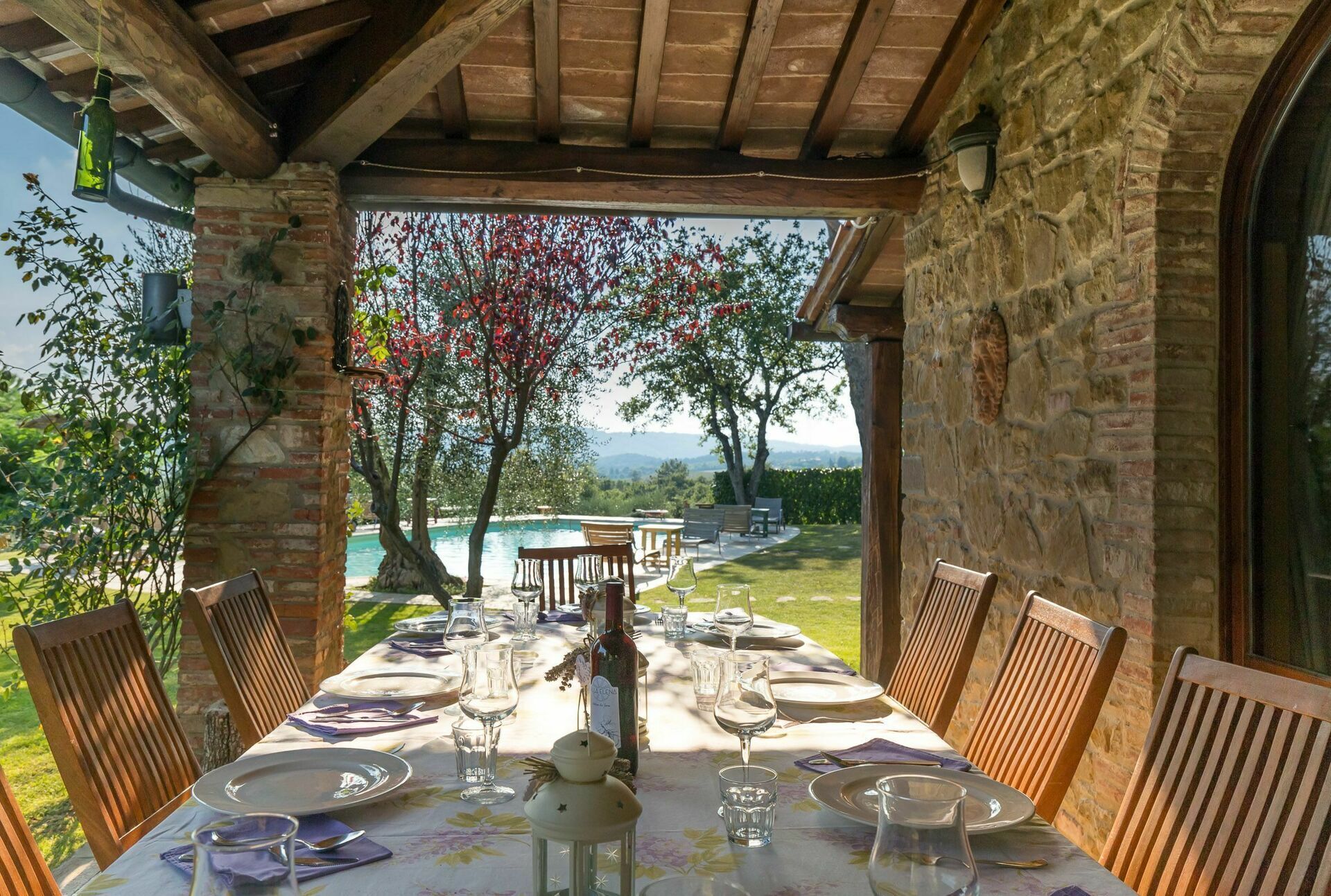 Zona pranzo all'aperto con tavolo imbandito per quattro persone, con vista su piscina e giardino.