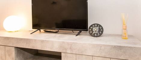 A flat-screen TV on a marble countertop with a round wall clock and a bottle of reed diffuser candles.