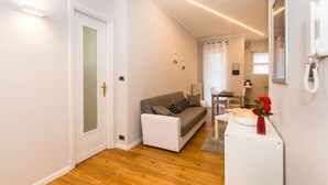 A modern living room with a sofa, a small table with a vase of flowers, and a kitchen area in the background. - GuestHost - Very new apartment equipped with large terrace and totally renovated, able to comfortably accommodate up to 3 people. The flat is located in the quiet Parella district, a very green and well served area: the MM Pozzo Strada stop is only 400m  (Torino)