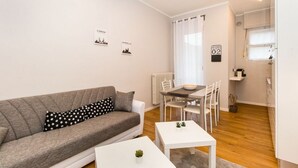 A modern living room with a grey sofa, white coffee table, and a dining area with a table and chairs. - GuestHost - Very new apartment equipped with large terrace and totally renovated, able to comfortably accommodate up to 3 people. The flat is located in the quiet Parella district, a very green and well served area: the MM Pozzo Strada stop is only 400m  (Torino)