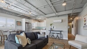 A spacious living area with a dark grey sofa, a white armchair, and a coffee table. A kitchen with white cabinets and a dining area with a small table and chairs are visible in the background.