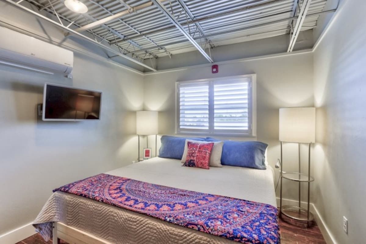 A bedroom with a bed, two bedside lamps, a window with blinds, and an air conditioning unit on the wall.