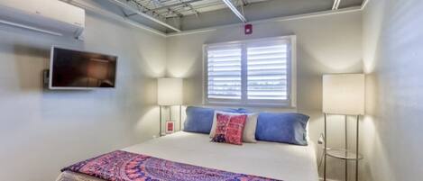 A bedroom with a bed, two bedside lamps, a window with blinds, and an air conditioning unit on the wall.