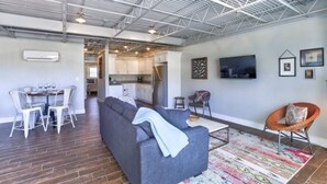 A modern living room with a grey sofa, a dining table with chairs, a kitchen area, and a TV mounted on the wall.