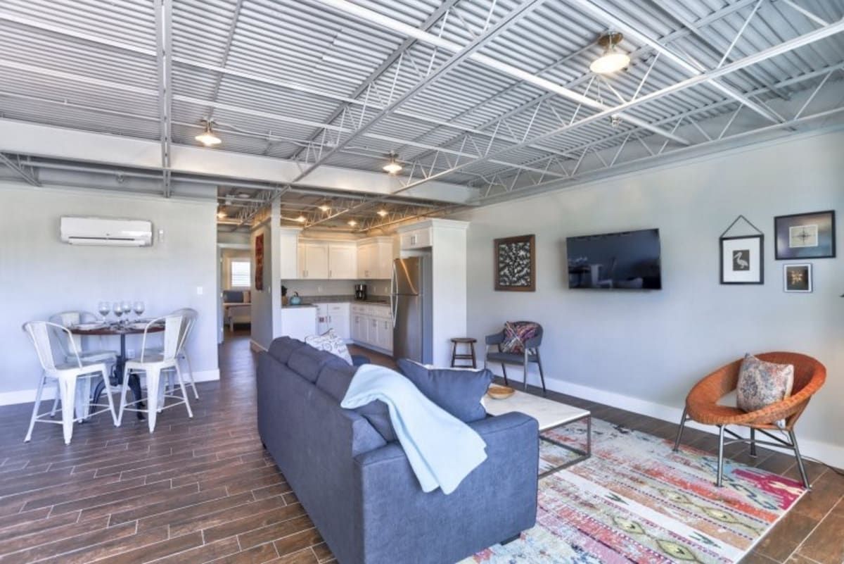 A modern living space with a grey sofa, a dining area with chairs, a kitchen in the background, and a flat-screen TV mounted on the wall.