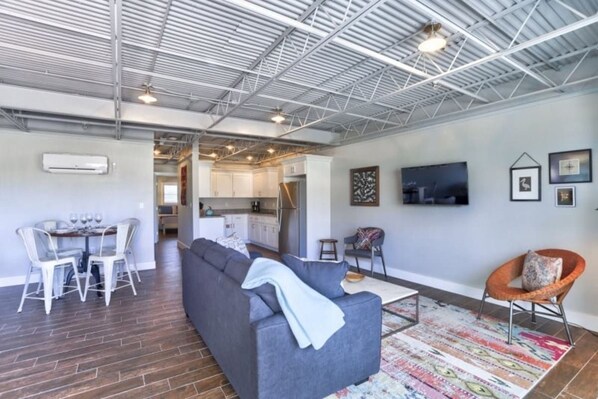 A modern living space with a grey sofa, a dining area with chairs, a kitchen in the background, and a flat-screen TV mounted on the wall.