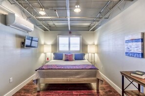 A bedroom with a bed, two bedside lamps, a television on the wall, and a window with blinds.