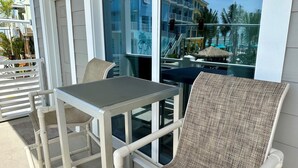 A balcony with a table and two wicker chairs, overlooking a pool area with umbrellas and a building in the background.