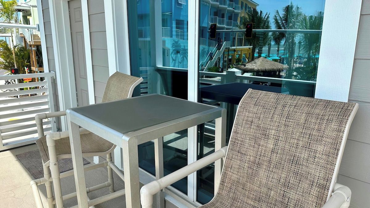 Varanda com mesa e duas cadeiras de vime, com vista para área de resort com palmeiras e piscina.