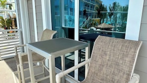 Un balcón con una mesa y dos sillas de mimbre, con vistas a una zona turística con palmeras y una piscina.