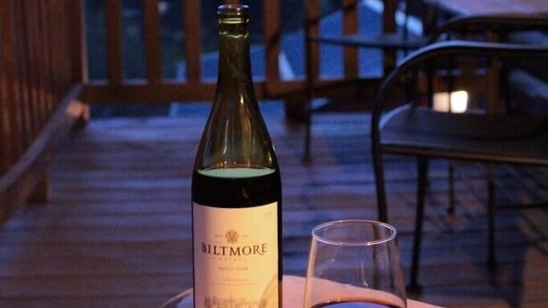 A bottle of Biltmore wine and a glass on a wooden table on a balcony with a view of mountains. - Turkey Trot- New Cabin in Black Mountain w/ views & discount Biltmore tickets! (Black Mountain)