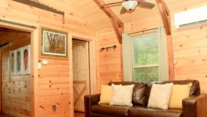 A wooden cabin interior with a brown leather sofa, wooden walls, a ceiling fan, and a window with a view of greenery. - Turkey Trot- New Cabin in Black Mountain w/ views & discount Biltmore tickets! (Black Mountain)