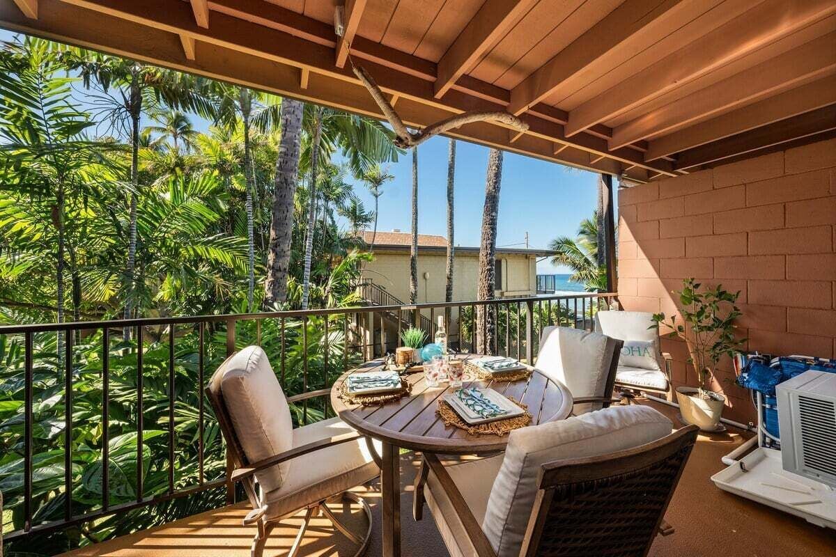 A covered outdoor seating area with a table set for two, surrounded by chairs, overlooking a tropical garden with palm trees.