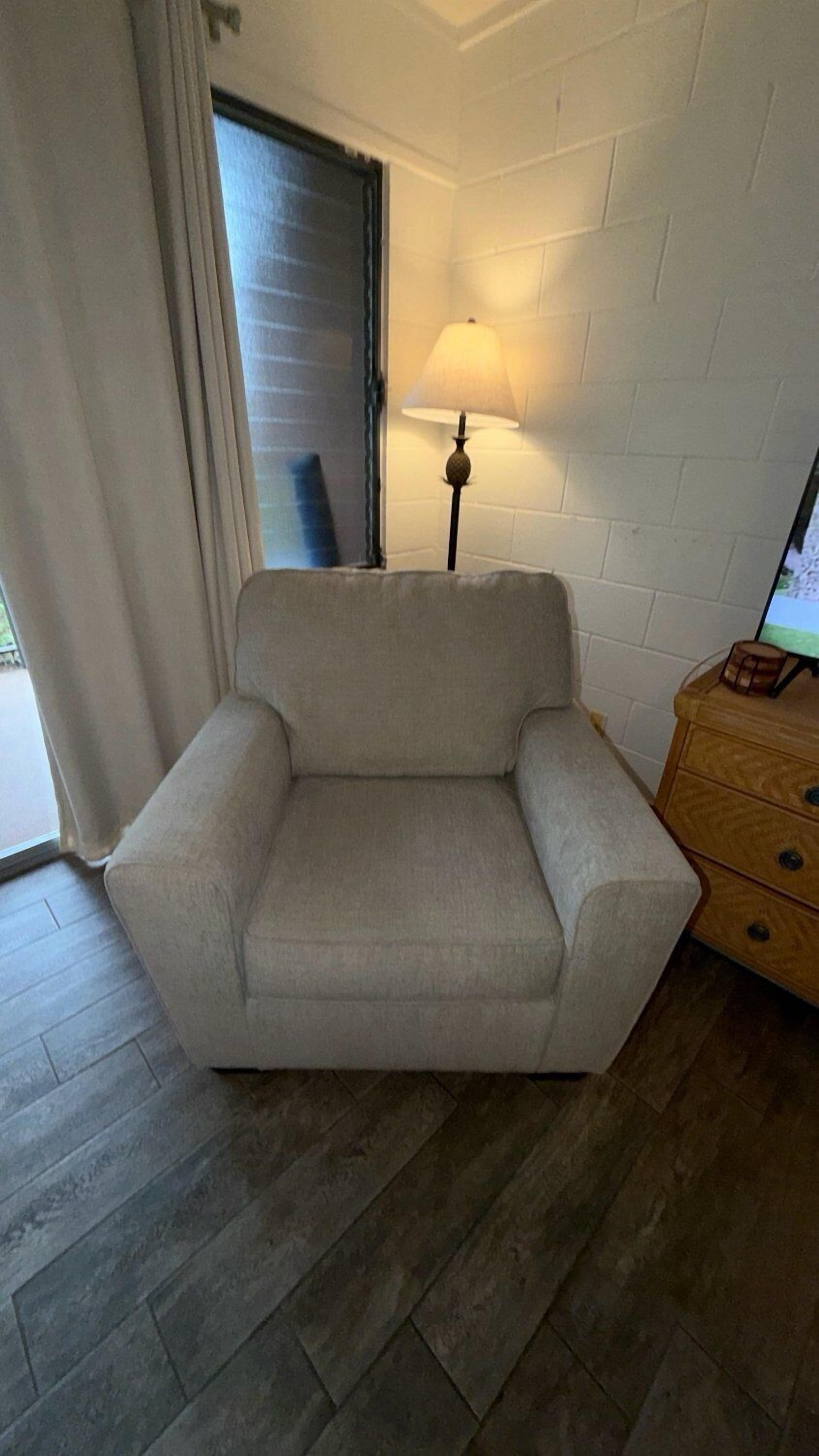 Une pièce avec un fauteuil beige, une commode en bois, une lampe et une fenêtre avec des rideaux.