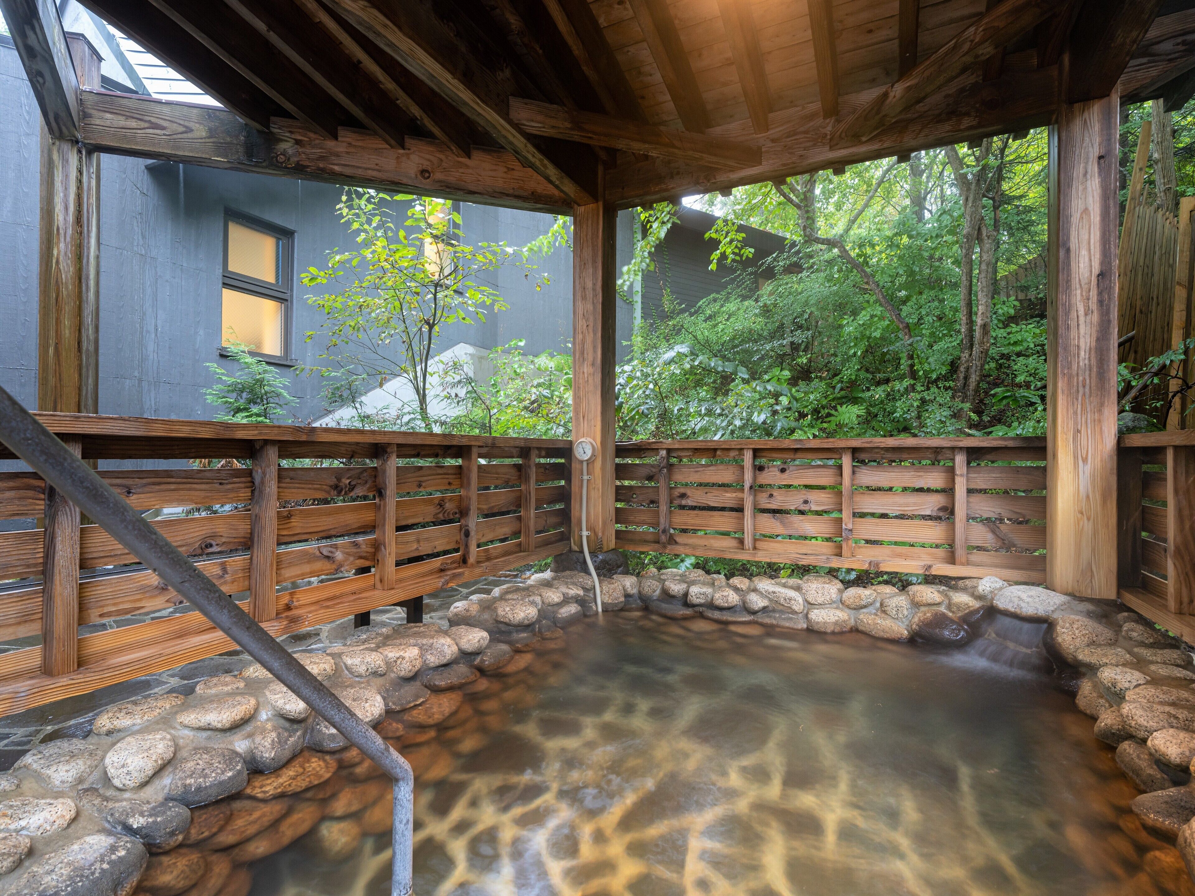 A wooden deck with a stone-lined hot spring, surrounded by a wooden fence and a view of a forested area.
