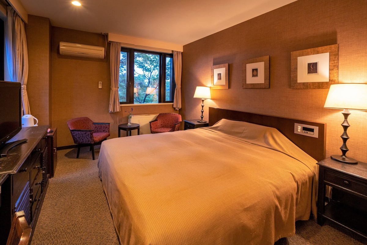 Een hotelkamer met een groot bed, twee fauteuils, een bureau met lamp en een raam met gordijnen.