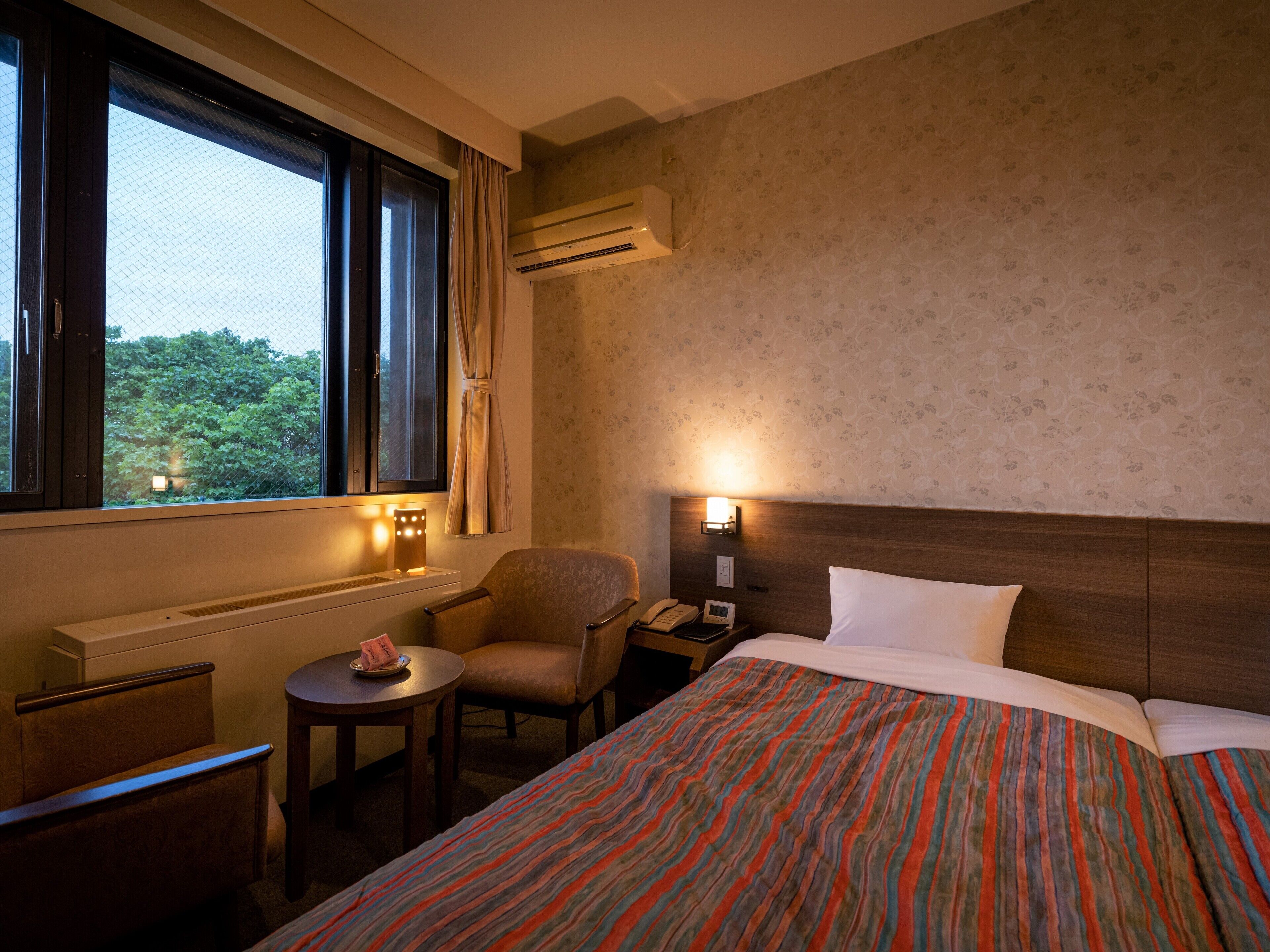 Habitación de hotel con cama, una silla, una mesita, aire acondicionado y ventana con cortinas.