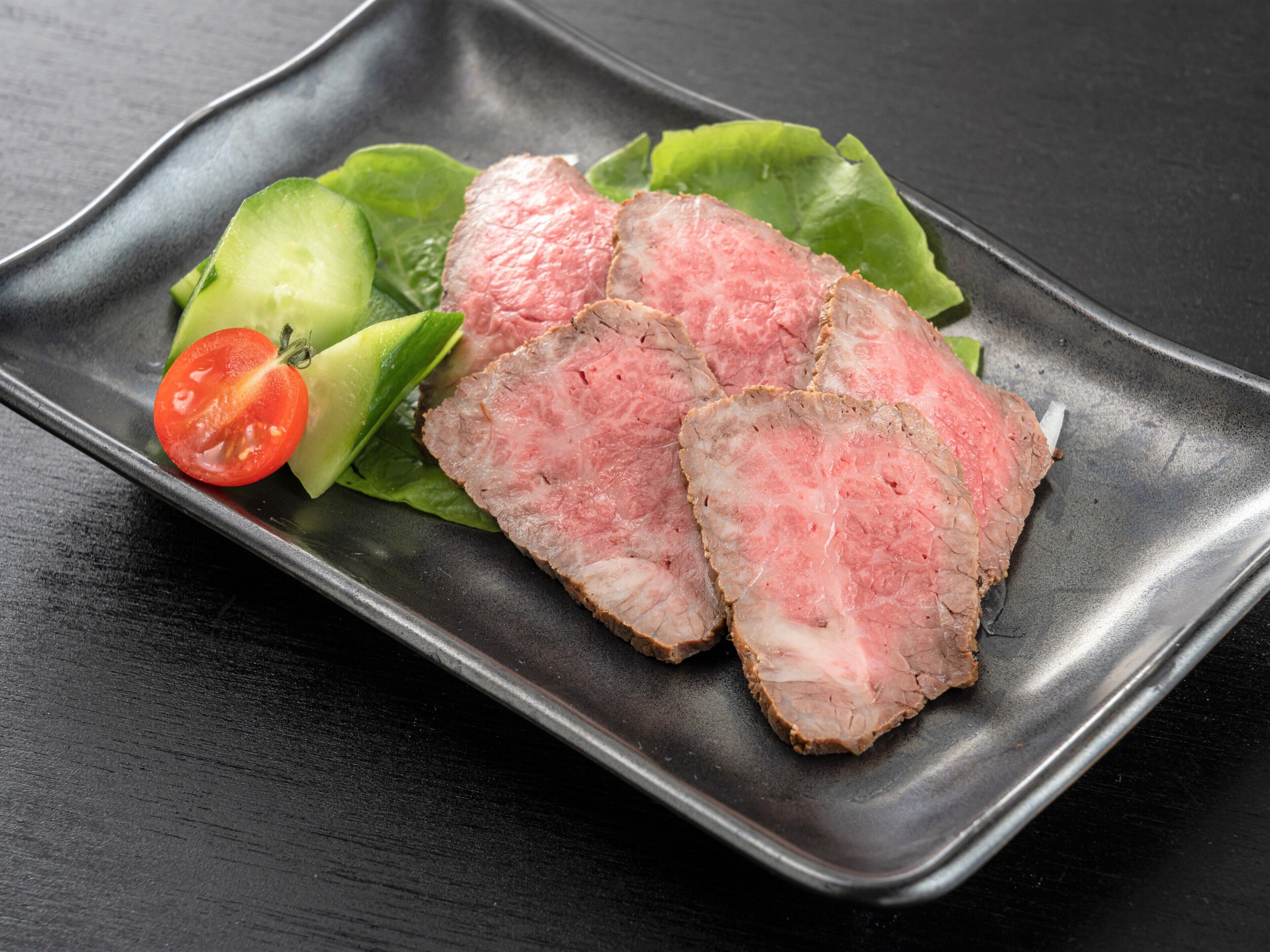 A plate of sliced meat with lettuce and a cherry tomato on a black plate.