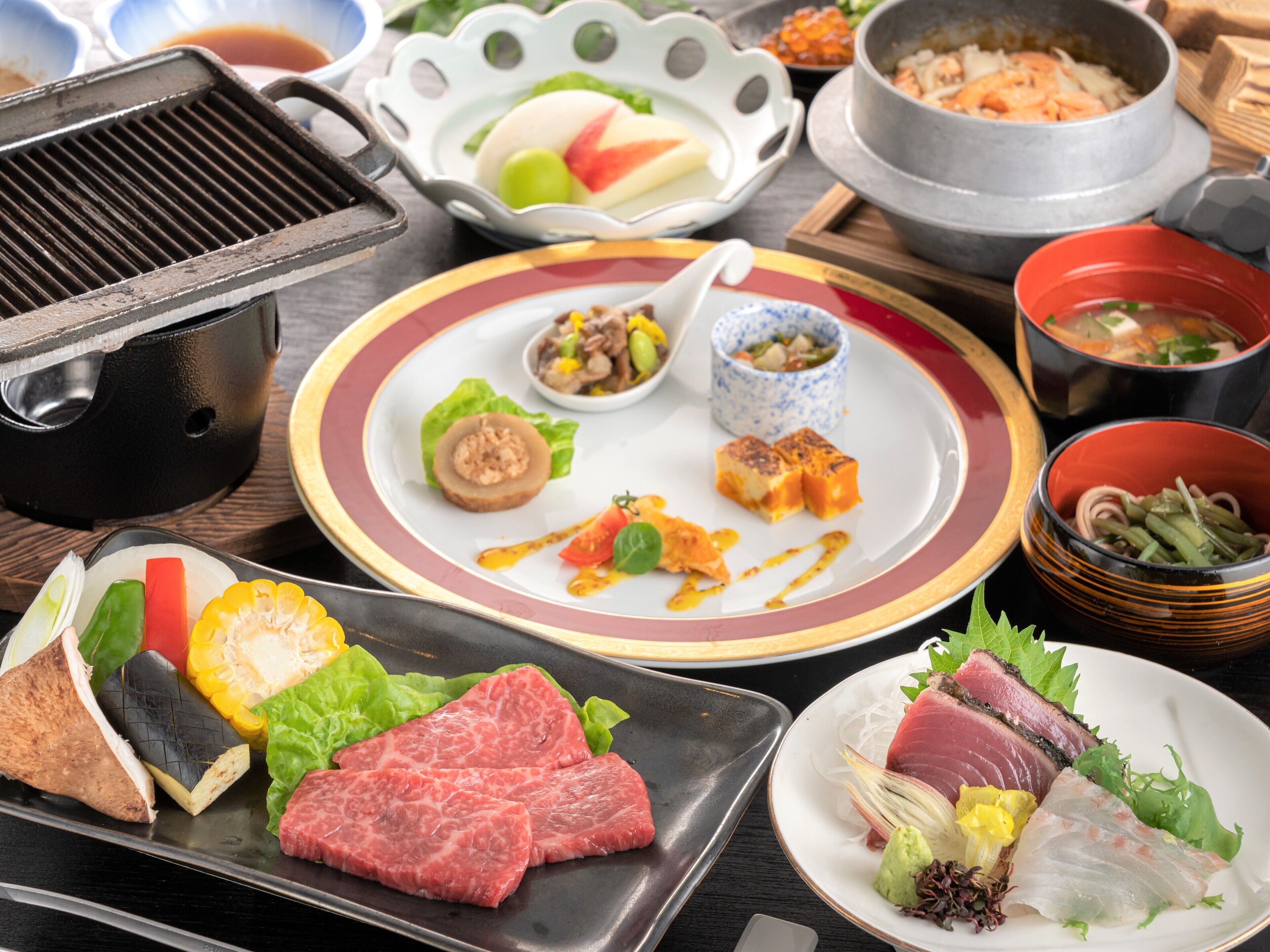 A variety of dishes including raw beef, vegetables, and seafood, arranged on a table with a grill and bowls of soup.