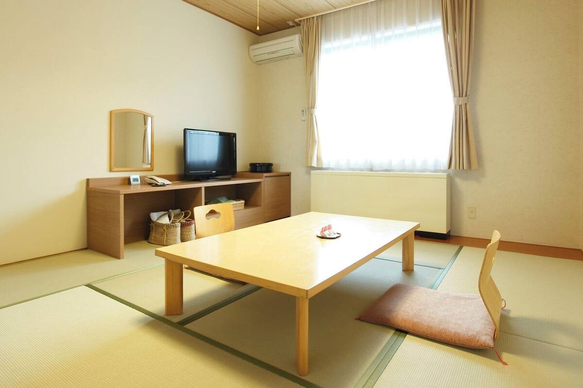 Een kamer met tatami vloerbedekking, een houten tafel, een televisie, een bureau met spiegel en een raam met gordijnen.