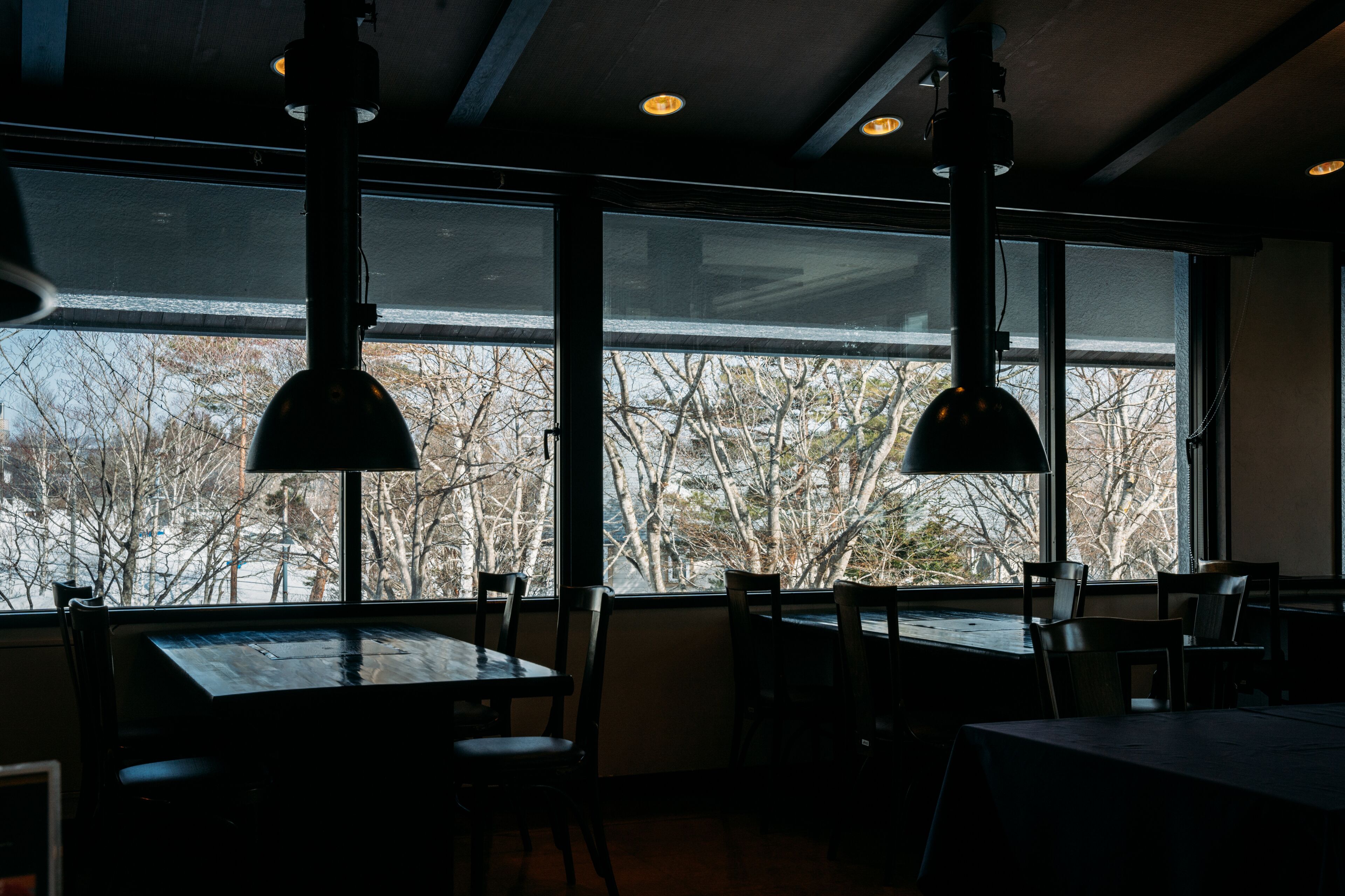 Interior de restaurante con amplias ventanas, mesas, sillas e iluminación colgante.