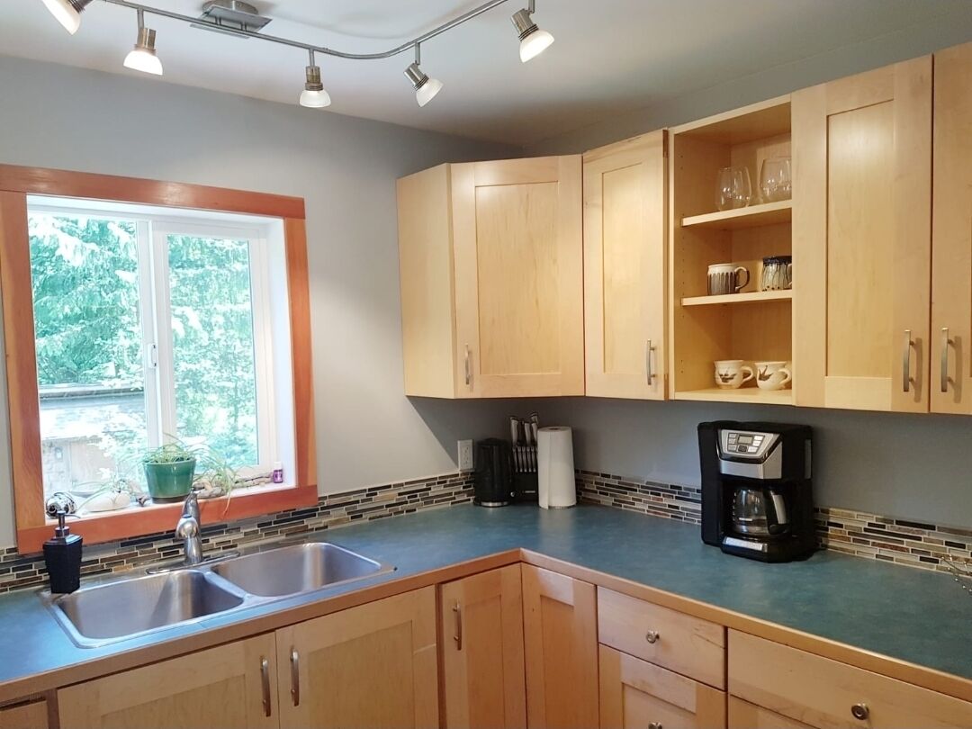 A kitchen with wooden cabinets, a double sink, a coffee maker, and a window with a view of greenery.
