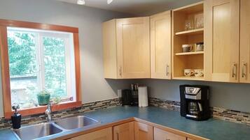 A kitchen with wooden cabinets, a double sink, a coffee maker, and a window with a view of greenery.