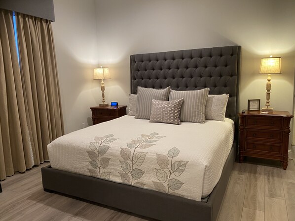 A neatly made bed with a tufted headboard, two bedside tables with lamps, and a large window with curtains. - Remodeled 3 BR 2BA condo steps to the beach! Sunset views and private garage. (Panama City Beach)