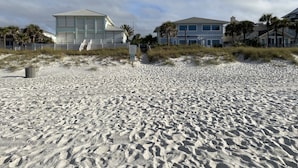 Uma praia arenosa com um edifício branco, com varanda e escadaria que dá para a praia.