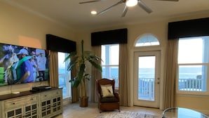 Sala de estar com televisão, planta, cadeira, mesa de vidro e vista para o oceano através de grandes janelas.