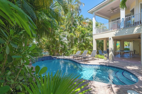 A swimming pool surrounded by lush greenery, with a covered patio area and lounge chairs.