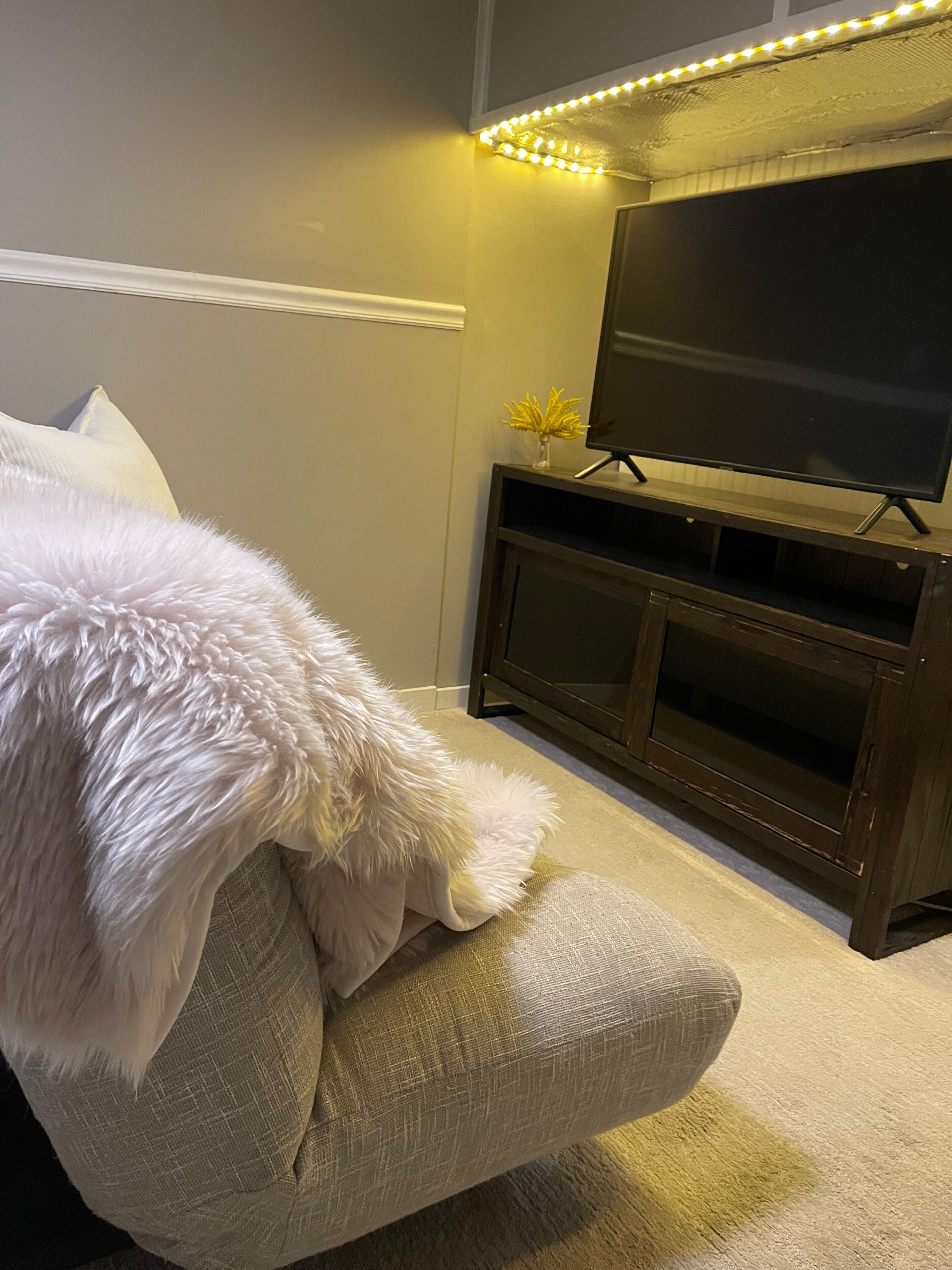 A room with a large flat-screen TV on a wooden stand, a beige sofa with a white furry throw, and a small plant on the stand.