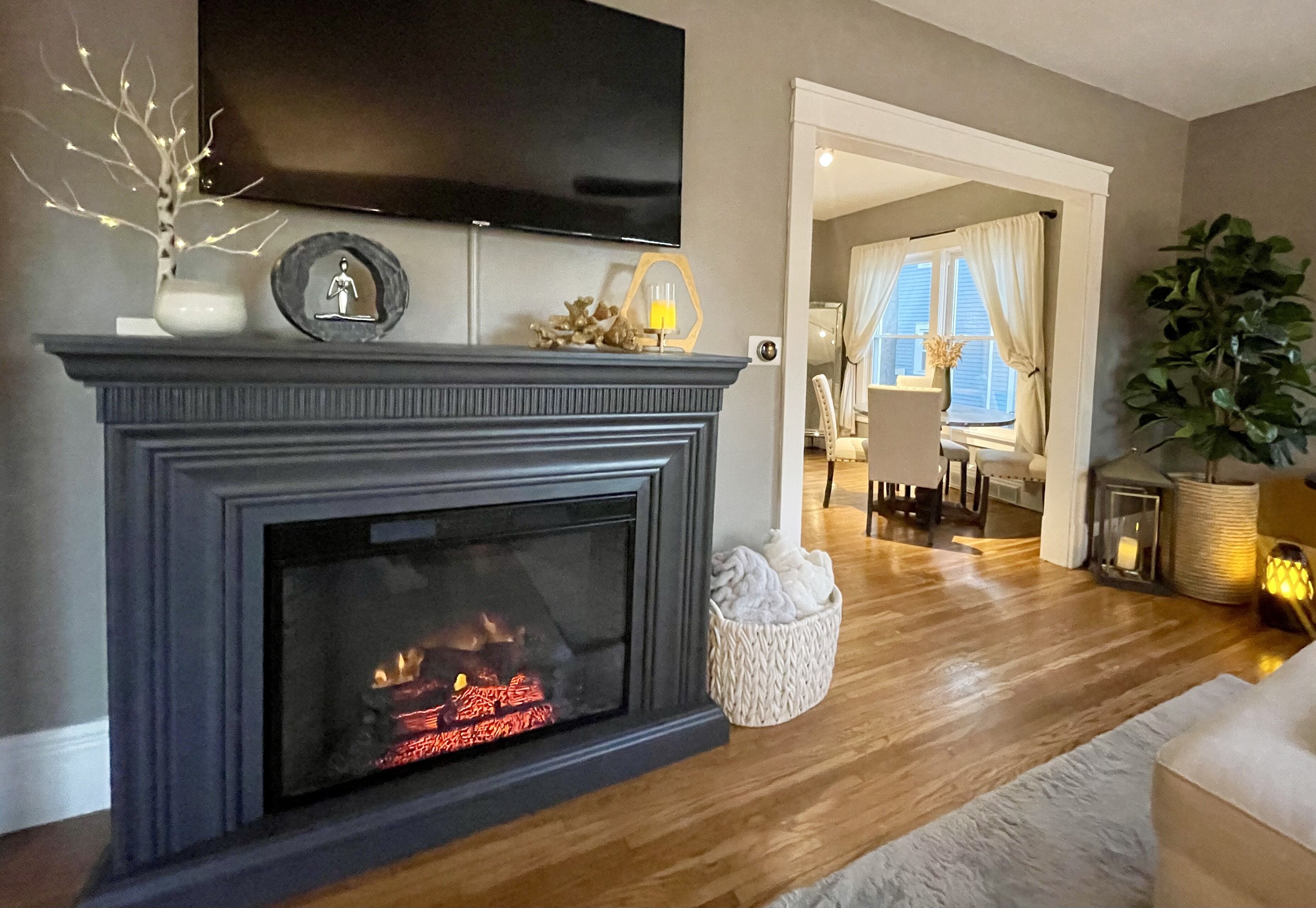 A living room with a fireplace, a flat-screen TV mounted above it, and a cozy seating area with a rug and a plant.