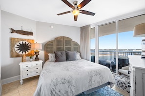 A bedroom with a large bed, a ceiling fan, a nightstand with a lamp, a fish decoration, and a view of the sea through a sliding door.