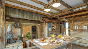 Un coin repas rustique avec une table en bois, un sol en pierre et une décoration champêtre comprenant un balai et une horloge. La table est mise pour le petit-déjeuner.
