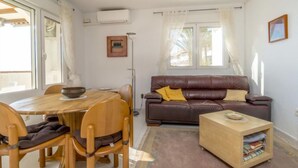A living room with a brown leather sofa, a wooden dining table with chairs, a small wooden side table, and a window with curtains.