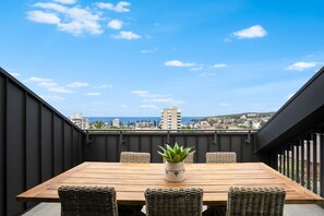 Ein Holztisch mit Korbstühlen und einem Blumentopf auf einem Balkon mit Blick auf Stadt und Meer.