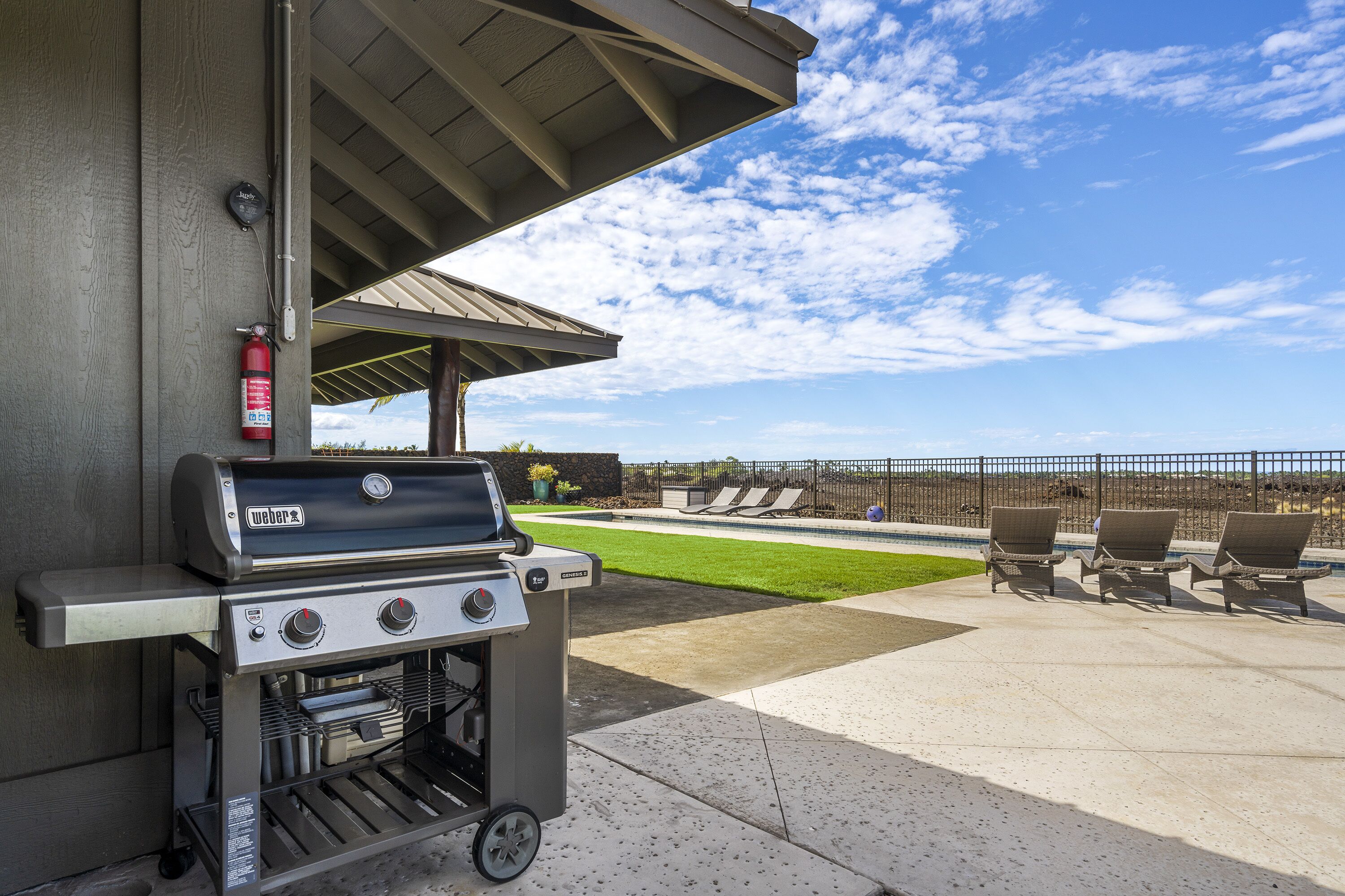 Un área de patio con una parrilla Weber Genesis II, mobiliario exterior y vista a una piscina y jardines.