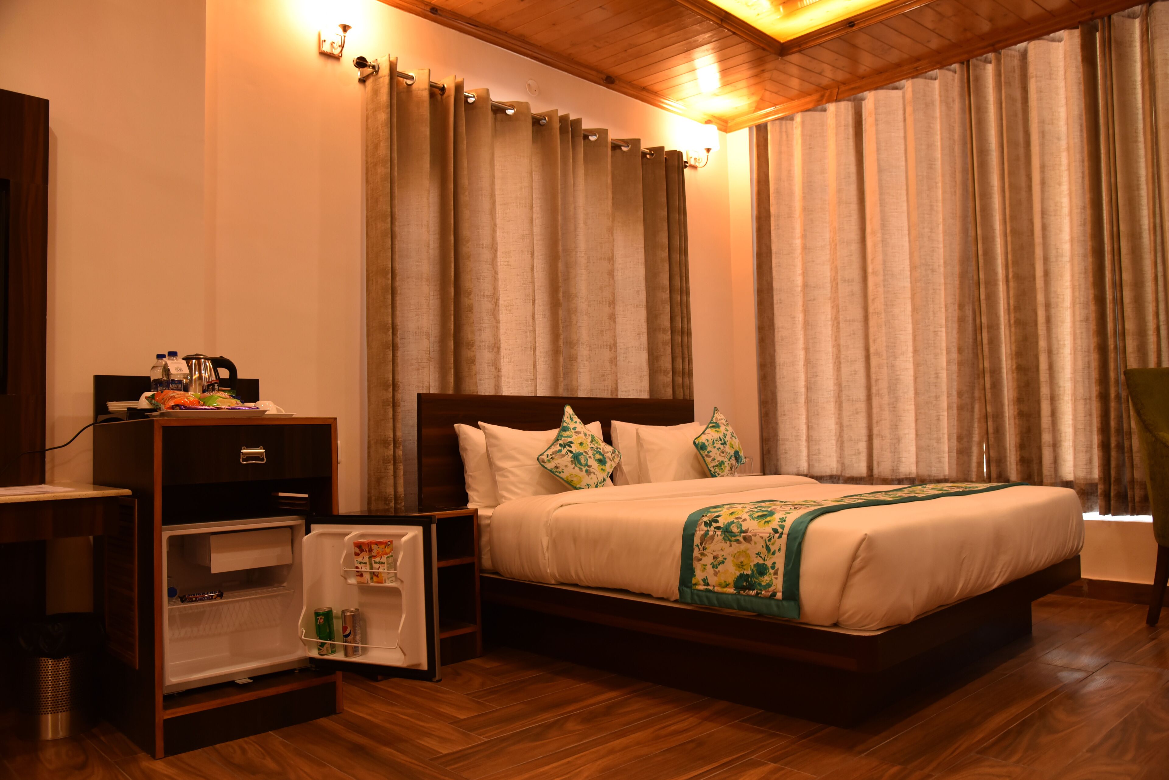 A hotel room with a wooden bed, a small refrigerator, a bedside table, and curtains.